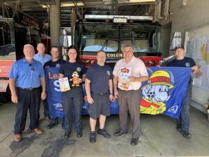 Male and female firefighter stand in from of a fire engine as they accept fire prevention week educations books from local insurance agents. Who firefighters are holding up a blue banner in the background that has a popular fire dog, sparky, on the banner. The fire engine towers over those standing in front. All are smiling and happy to promote fire prevention as they stand in the garage of the fire station 1.