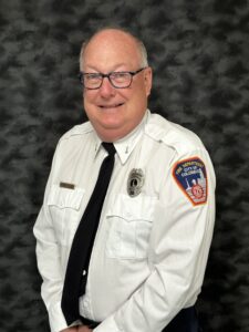 Lt. Jim Miller Stands with this arms crossed wearing his Columbus Fire Department Uniform. Miller is smiling at the camera. A dark backdrop contrasts well with Miller's white uniform shirt , which is adorned with his fire department badge and the Columbus Fire Department patch on his dress shirt. 