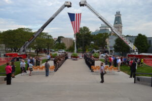 An American flag is hoisted above the Columbus City Hall Plaza with a crowd of community members and public safety officers standing at attention in anticipation for the start of the annual 9-11 remembrance ceremony. Public safety officers are dressed in their ceremonial uniforms.