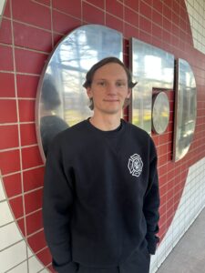 Drew Baldwin stands within Columbus Fire Station 1 wearing a blue CFD sweatshirt. The background included a large aluminum CFD sign sounded by red brick. Drew, with a closed mouth smile, stands with his hands in his pants pockets eager to begin his work as a fire inspector 