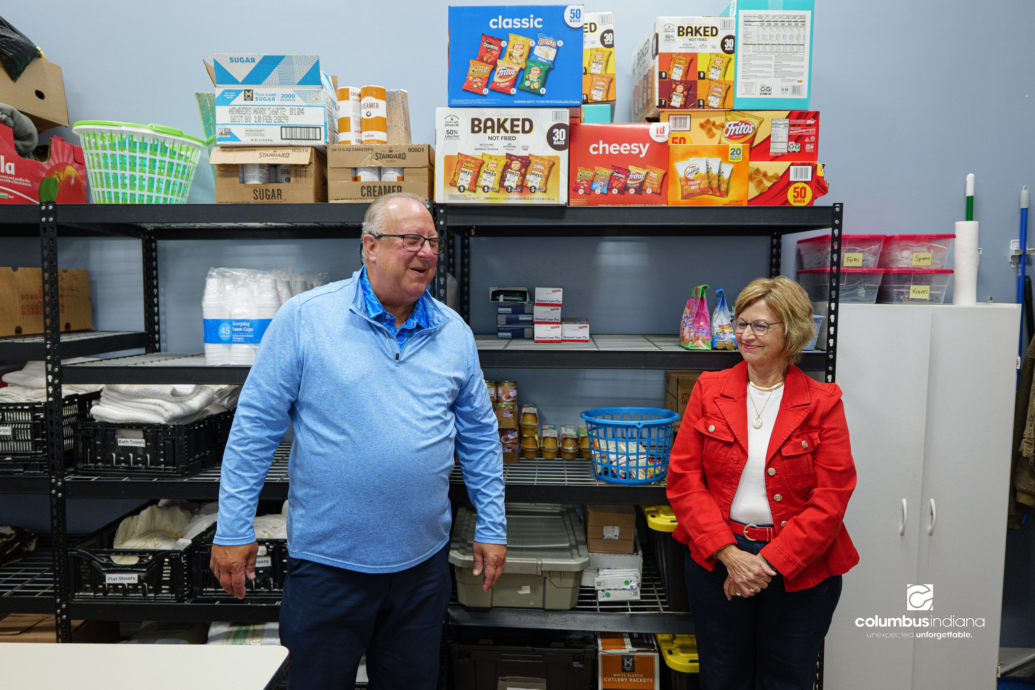 Two people stand in front of a shelf full of food talking.