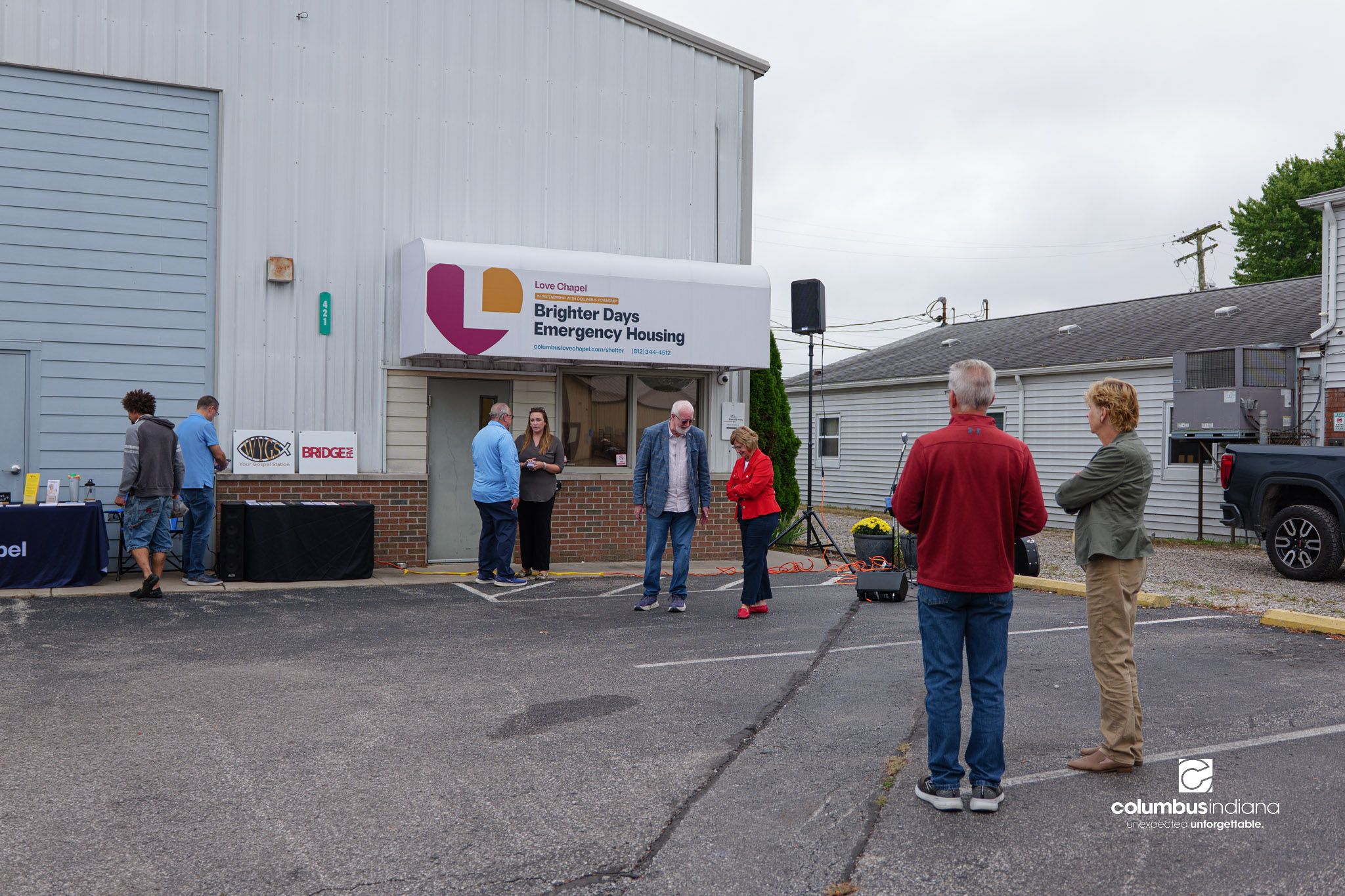 A group of people stand outside a building. The sky is overcast and gray. A public address system is set up for speeches.