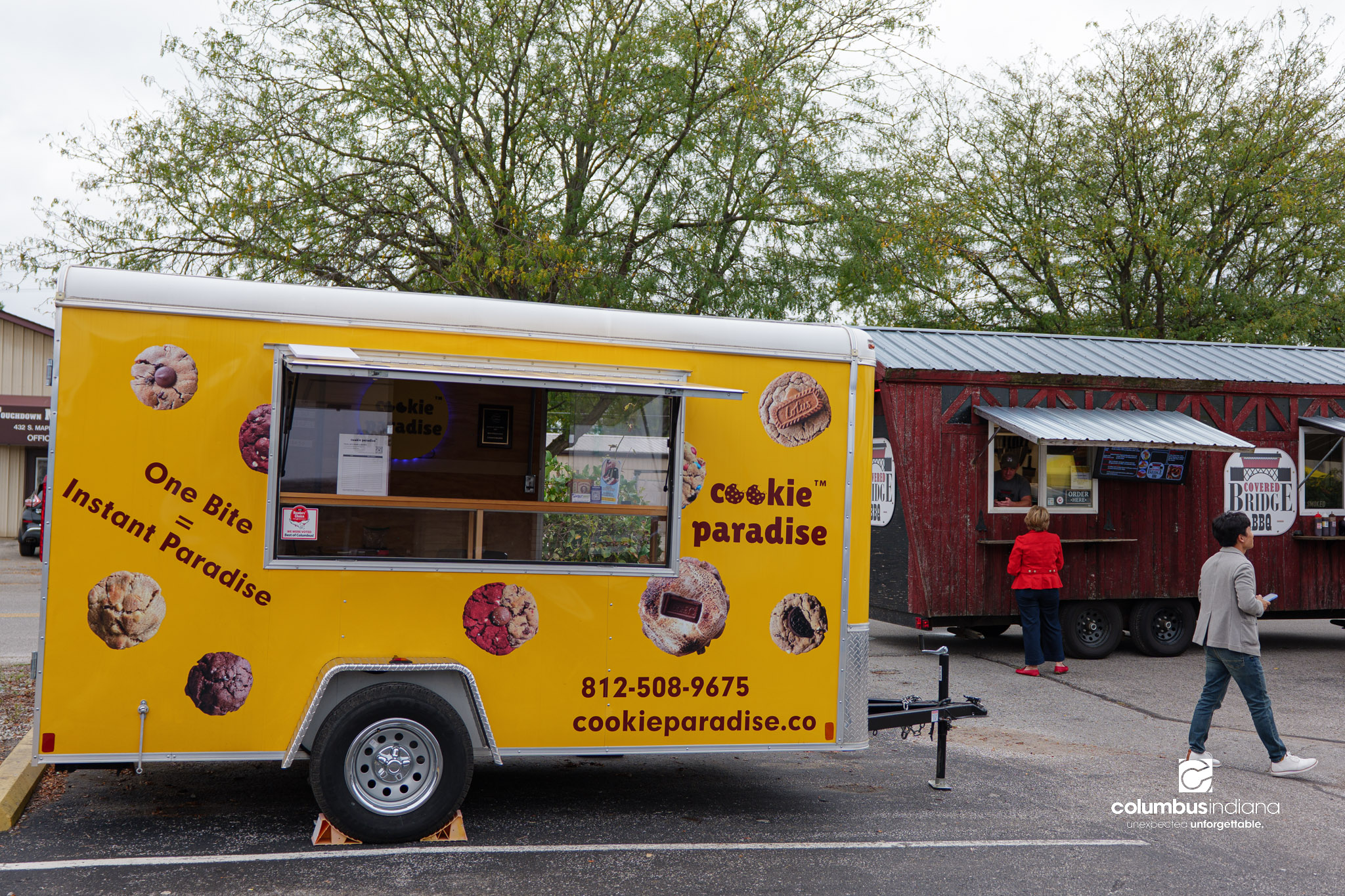 Food trucks are parked on a street. The one on the left is red and looks like a covered bridge. The one on the right is yellow with graphics of cookies.