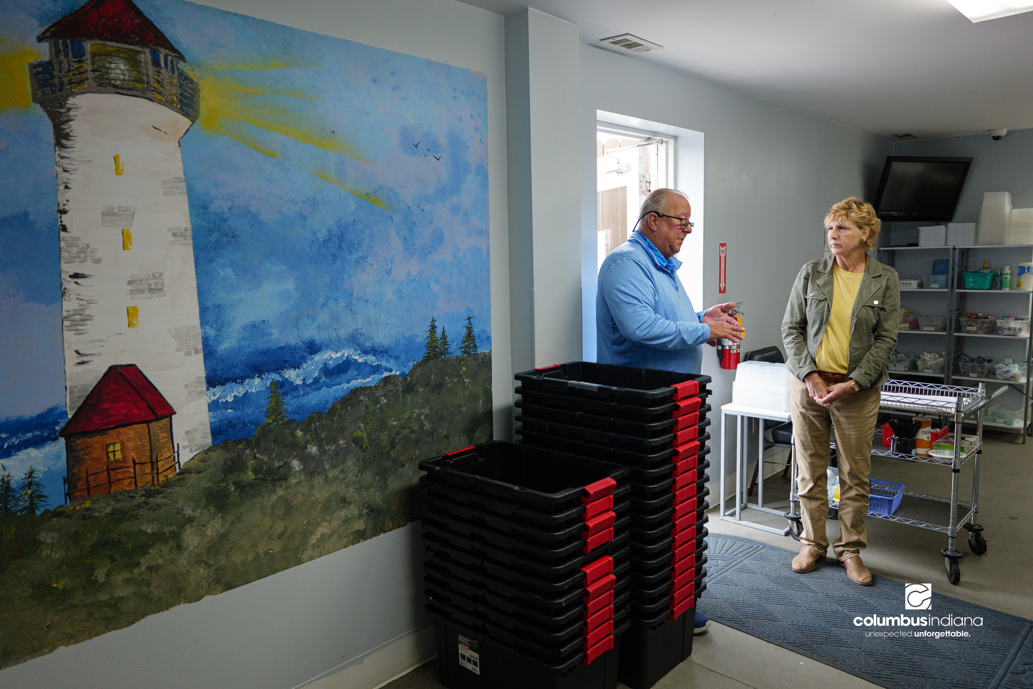 Two people talk in a room. There are crates stacked up on a wall and the wall features a mural of a lighthouse.