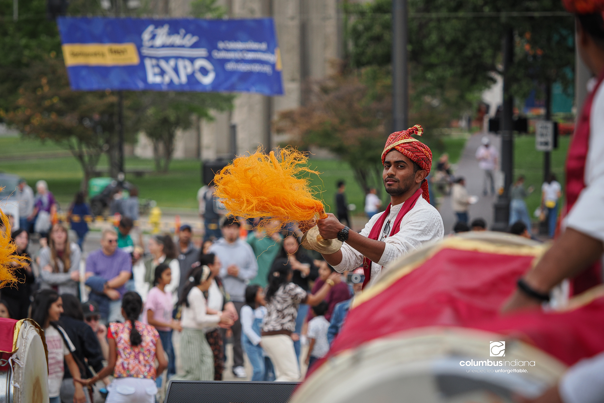 A group of Indian musicians put on a musical performance with percussion istruments of various sizes. They are dressed in white clothes with colorful red and gold headdresses and scarves.